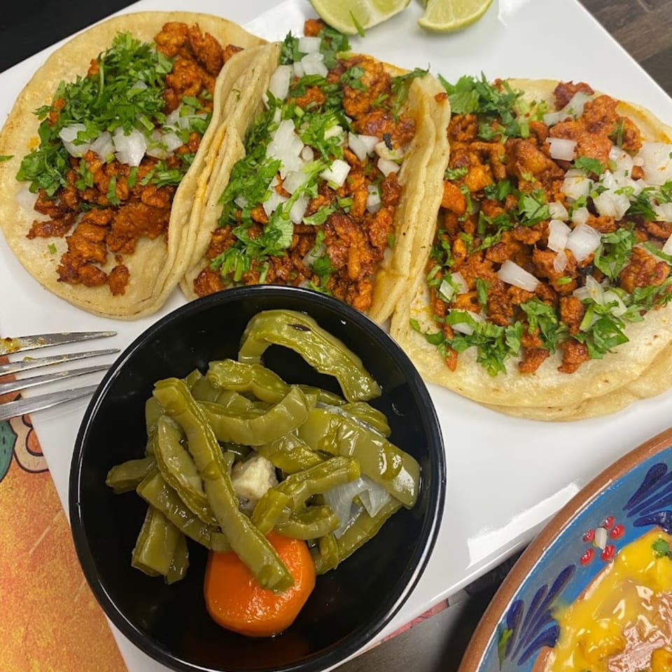Three al pastor tacos with cilantro and onions, served with a side of nopales and a lime wedge.