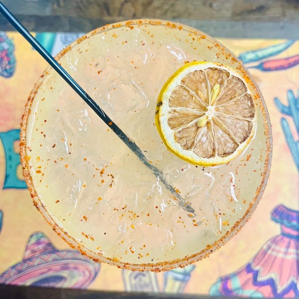 Overhead view of a margarita with a dried lemon slice, black straw, and chili-salted rim.