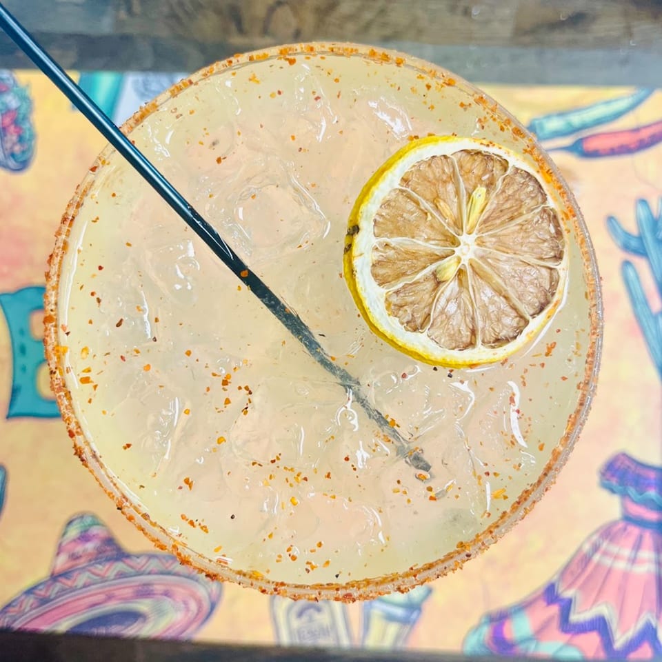 Overhead view of a margarita with a dried lemon slice, black straw, and chili-salted rim.