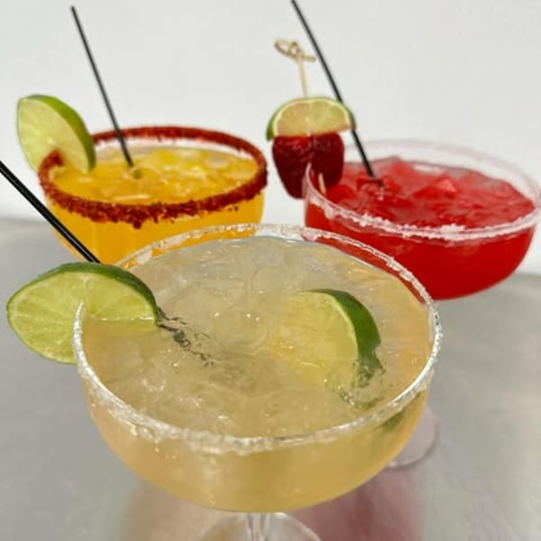 Three colorful margaritas, one yellow, one red, and one clear, garnished with lime and strawberry.