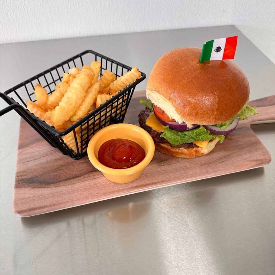 A burger with a Mexican flag pick, crinkle-cut fries in a basket, and ketchup on a wooden board.