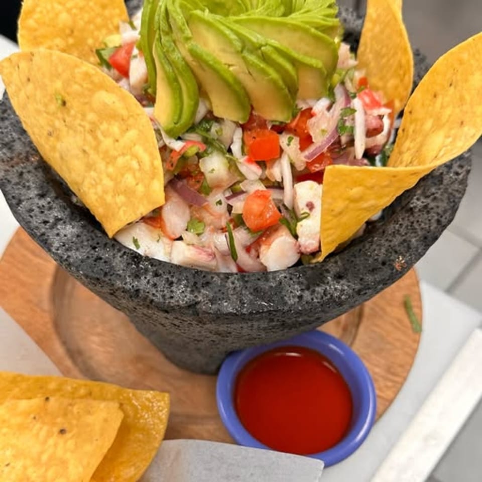 Mexican ceviche in a stone bowl with avocado slices, tortilla chips, and a side of red sauce.