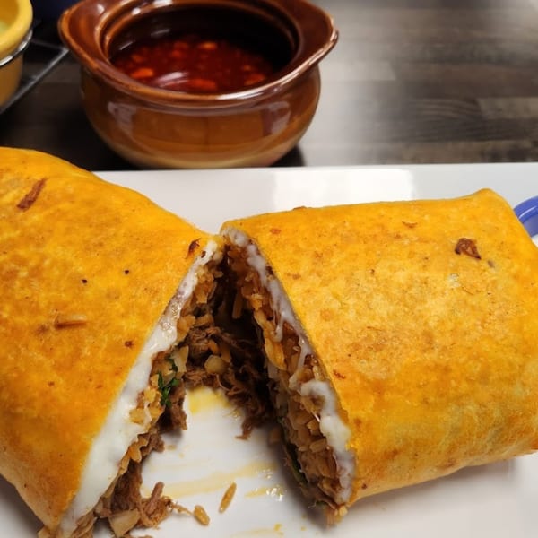 A large, golden-brown burrito cut in half, revealing shredded meat, rice, and white sauce, with a side of red salsa.