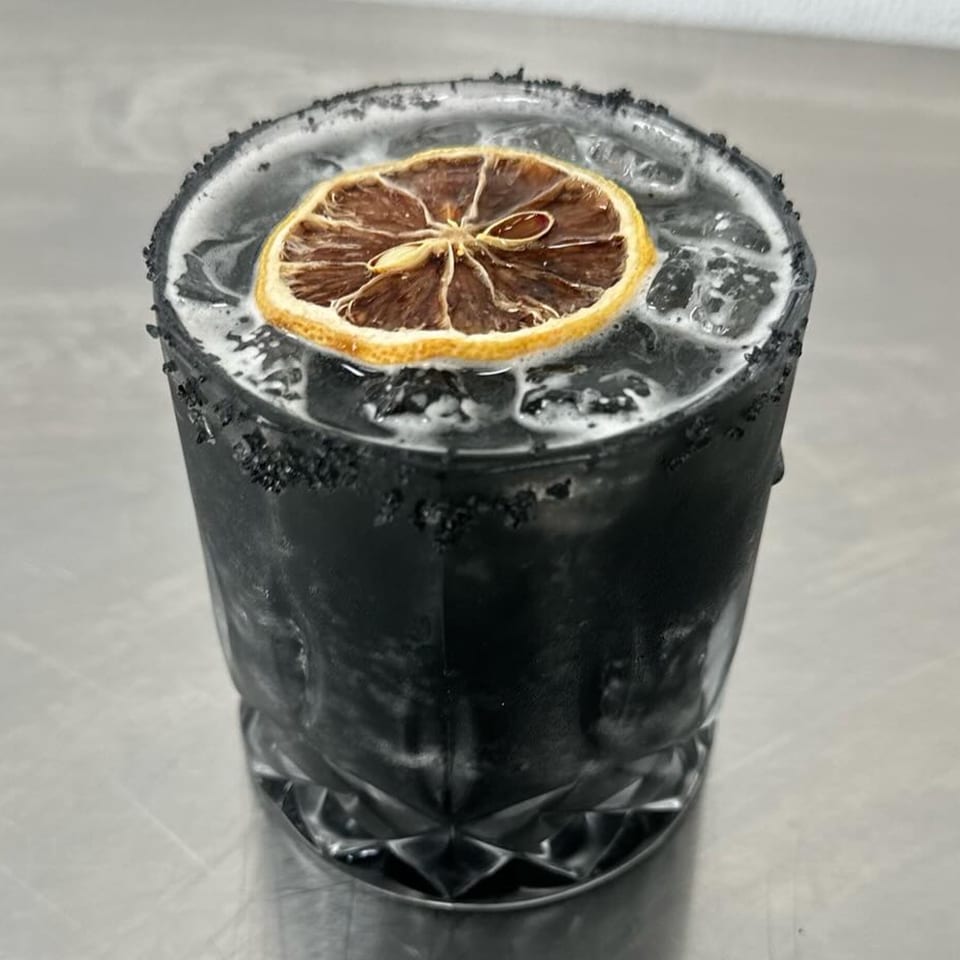 A dark cocktail with a dried orange slice and a black sugar rim, on a metal surface.