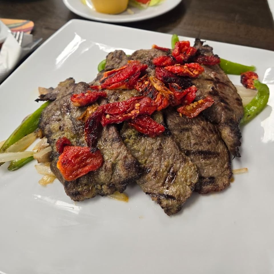 Grilled steak topped with sun-dried tomatoes, green peppers, and onions on a white plate.